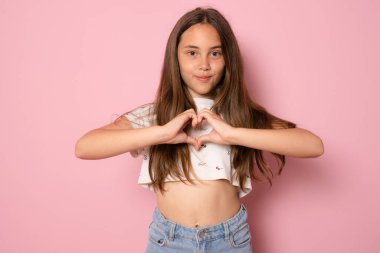Pembe arka planda izole edilmiş tişört giyerek elleriyle kalp formu oluşturan güzel bir genç kızın portresi.