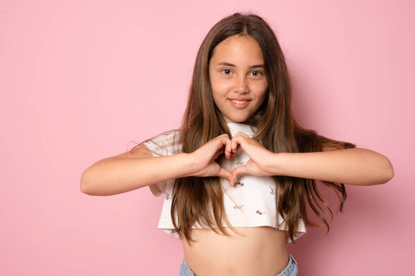 Портрет милой девочки-подростка, делающей форму сердца с улыбкой на руках на розовом фоне в футболке