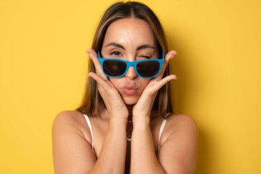 Gülümseyen esmer kadının yakın portresi. Güneş gözlüklü genç kadın. Sarı arka planda izole edilmiş bir öpücük yolluyor..