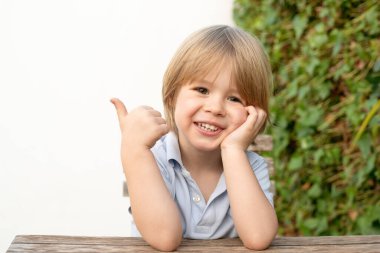 Masada oturan sevimli sarı saçlı çocuk ve baş parmağıyla gülümsüyor. Arka planda taze yeşil yapraklar..