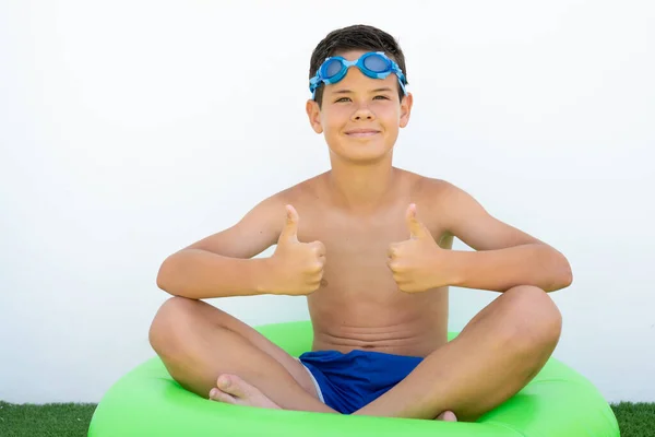 Happy boy sitting in swim ring on grass showing thumbs up outdoors.