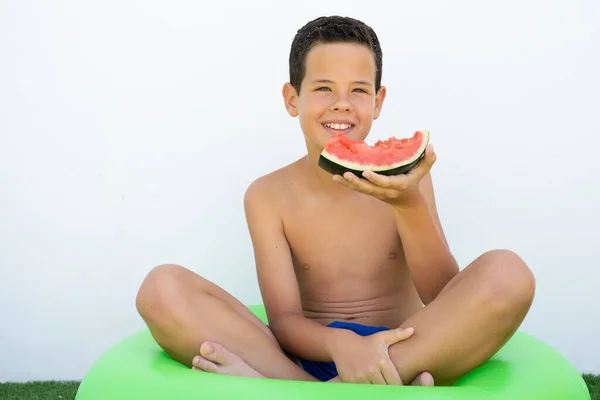 Happy boy sitting in swim ring on grass eating watermelon outdoors.