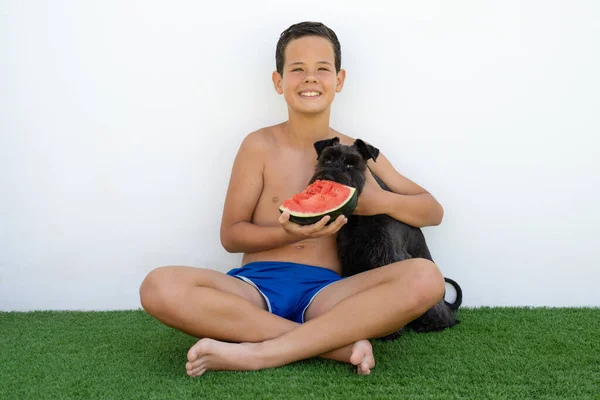 Smiling boy in summer clothing sitting on the grass hugging his dog eating watermelon.