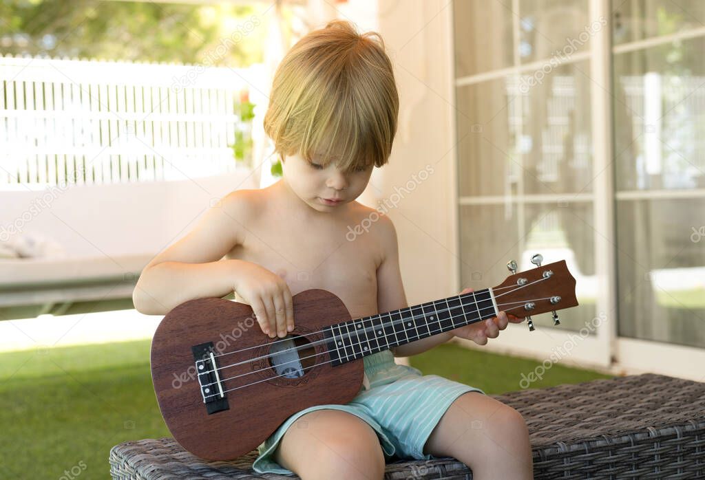 Little happy boy plays his guitar or ukulele, sitting in the terrace outdoors