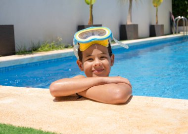 Mutlu genç bir çocuk yüzme havuzunun kenarında, sarı gözlük takıyor.