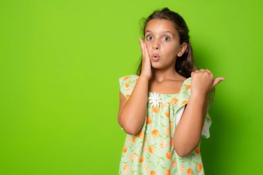 Şok olmuş, tatlı, esmer, küçük bir kız çocuğu, yüzü şaşkın, sağa dön, sağ fotokopi alanını şaşırt, çeneni indir hayret et, nutku tutmayan ve hayrete düşmüş harika bir reklam, yeşil arka planda dur, eğlen