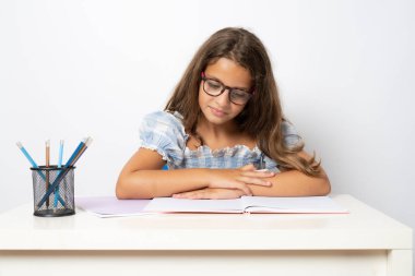 Portrait of positive school girl sitting behind desk reading a book isolated on white background