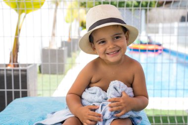 Hasır şapka takmış, açık havuzun yanında oturan sevimli, gülümseyen küçük çocuk.