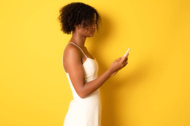 Side view of smiling afro american young woman using smart phone over yellow background