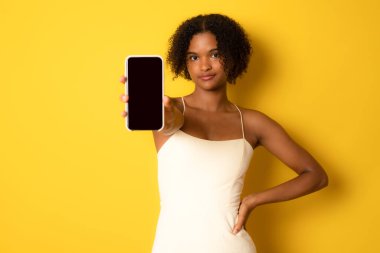 Portrait of a smiling young african woman holding blank screen mobile phone over yellow background