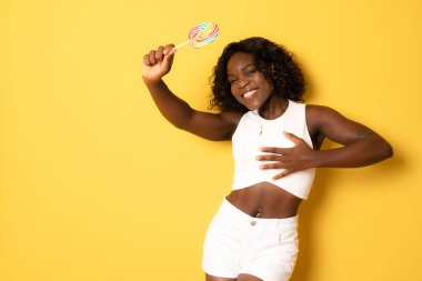 Kıvırcık saçlı, güzel ve pozitif bir kadın elini göğsüne koyuyor. Gülümsüyor. Lolipop sarı arka planda izole edilmiş günlük kıyafetler giyiyor. Tatlıya düşkünlük