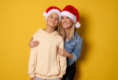 Orta yaşlı güzel bir anne ve yakışıklı oğlu Noel Baba şapkası takıyor. Sarıda izole edilmiş..