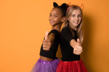 Siyah ve renkli cadılar bayramı kostümlü gülen çok uluslu kızların resmi turuncu duvarda başparmaklarını gösteriyor.