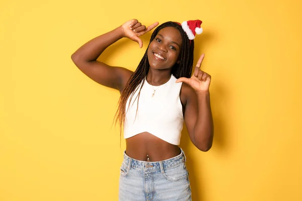 Portrait of beautiful african american female woman model wearing santa hat making frame gesture with fingers isolated over yellow background.