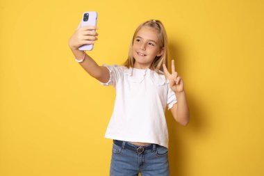 Beyaz tişört giyen küçük bir kızın portresi cep telefonunun kamerasına barış işareti ya da Ziyaretçi jesti yapıyor. Selfie ya da video görüşmesi yapmak için poz veriyor. Sarı arka planda kapalı stüdyo çekimleri.