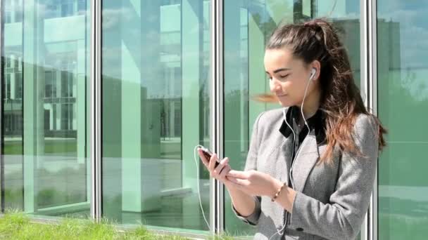 Bussines woman listens to music on headphones before bussines building — Stock Video