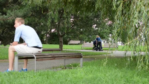 Мужчина сидит в парке на скамейке - женщина с ребенком в коляске на заднем плане — стоковое видео