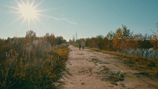 A falusi úton, melyen két gyermek jár, a nővér mutat valamit a fiatalabbnak a kék ég felé. Ragyogó nap, ősz — Stock videók