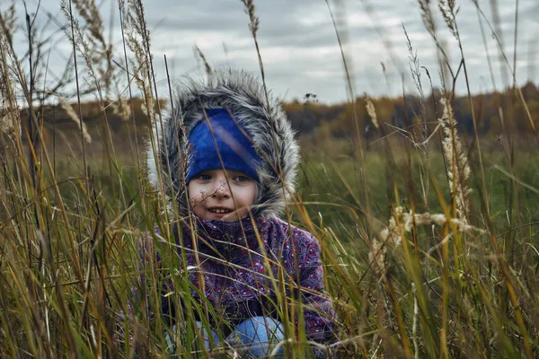 一个穿着紫色夹克和蓝色帽子的女孩坐在草地上 直直的看着 干枯的草叶 厚重的云彩在天空中 — 图库照片