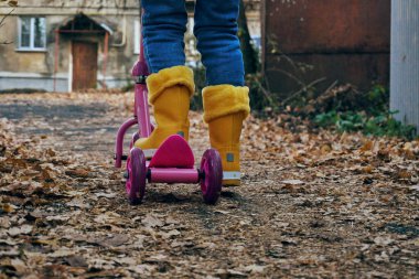 Sarı lastik çizmeli, pembe scooterlı bir kız. Sonbahar, yapraklar asfalt kaldırımda yatıyor..