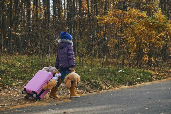 Осенью девочка в жёлтых резиновых сапогах выходит на дорогу, в руках у неё розовый чемодан, наполненный мягкими игрушками, а в другой руке медвежонок — стоковое фото
