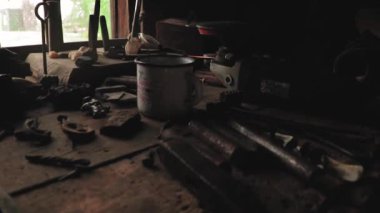 Demirci çalışma masası, beyaz demir bardak, ahşap bir masada demirci aletleri, demirciler için metal parçalar, pencerenin arkasından gelen doğal ışık.