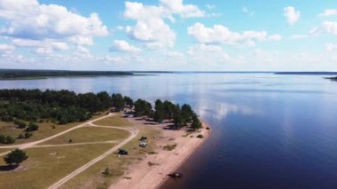 Mavi bir göl üzerinde pürüzsüz bir dron uçuşu ve yeşil ağaçlar ve çalılar, kumlu sahil ve yollar, sahildeki arabalar, açık hava eğlencesi, kamp, bulutlu mavi gökyüzü