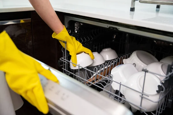 Close up hands of housewife wear yellow gloves loading dishwasher with clean dishes in the white kitchen.