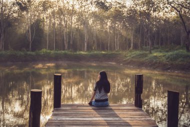 A sad woman sits floating on an old wooden bridge that juts out into a pond in nature. Lonely sad woman.