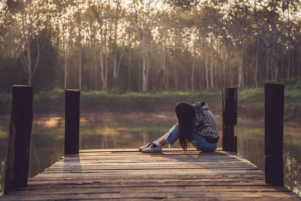 Crying woman hugging her knees on an old wooden bridge jutting out into a pond in nature. Lonely sad woman.