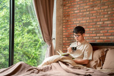 Evdeki yatak odasında elinde kahve fincanı olan bir adam kitap okuyor.. 