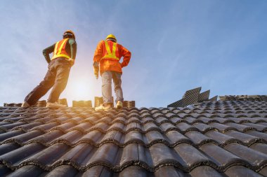 Koruyucu üniformalı iki Roofer işçisi, hava veya pnömatik çivi tabancası kullanarak ve beton çatı kiremitlerini yeni çatının üstüne monte ederek, inşaat halindeki konut kavramını kullanarak aşındırıyor..