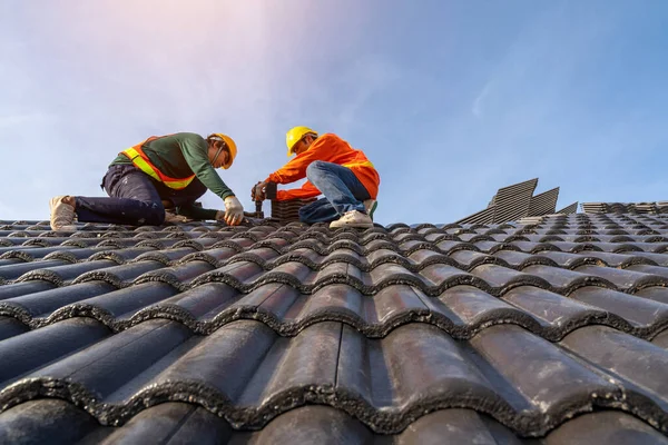 Teamwork of Asian roofer workers in protective uniform wear and gloves, using air or pneumatic nail gun and installing Concrete Roof Tiles on top of the new roof, building under construction.