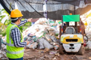 Asian  foreman working in recycling factory and holding tablet and looking at recycle waste factory with a forklift on the background. 