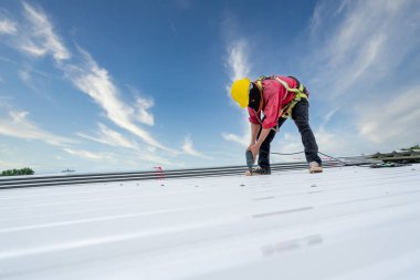 A Roofer working on roof structure of building on construction site, Roofer using electric drill nail gun and installing white Metal Sheet on top new roof. Safety body construction