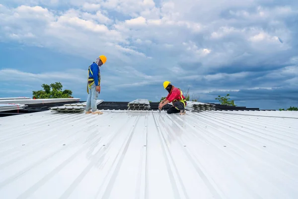 Roofer using electric drill nail gun installing Roof sheet under construction. Construction worker wearing safety harness belt during working on roof structure of building on construction site. 
