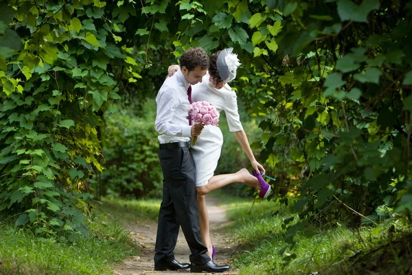 Wedding theme. The groom embraces the bride in a botanical garden. Bride adjusts her shoes. — Stock Photo, Image