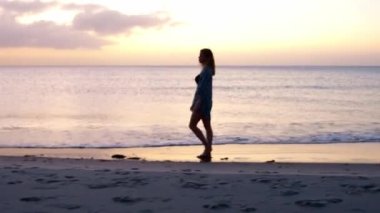 Bikinili genç bir kadın gün batımında sahilde yürüyor.