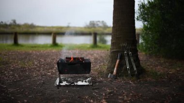 Kamp ateşi manzarası, nehrin yanında ateş çukuru, sağında ağaç ve çalılar var..