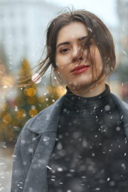 Güzel esmer kadın, kar yağışı sırasında Noel süsü verilmiş bir şekilde şehre yürürken şık bir palto giyiyor. Yakın çekim.