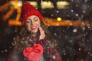 Memnun genç bayan kar yağarken kırmızı şapka takıyor ve elinde fiyonk var.