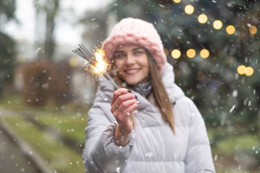 Göz kamaştırıcı gülümseyen kadın, kar yağarken yeni yıl ağacının yanında maytaplarla eğleniyor.