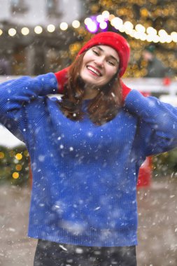 Güzel esmer kadın, kar yağarken Noel panayırında yürürken kırmızı şapka ve mavi kazak giyiyor.