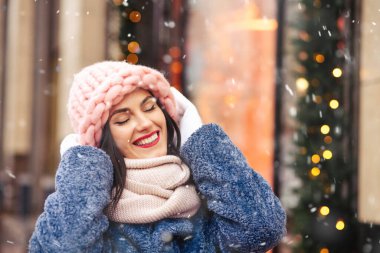 Neşeli esmer kadın, kar yağarken şehirde süslenmiş açık pembe bir şapka ve eşarp takıyor. Metin için boşluk