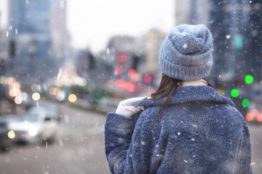 Kadın, karlı havada şehir trafiğine bakarken şık bir kışlık ceket ve şapka giyiyor. Metin için boşluk