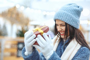 Kışlık ceketli, heyecanlı esmer kadın, kar yağışı sırasında Noel panayırında hediye kutusu tutuyor. Metin için boşluk