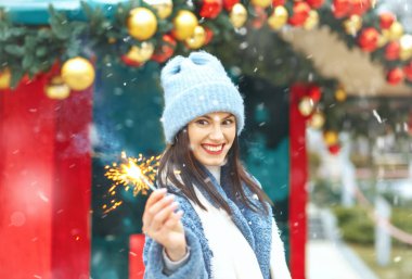 Şaşırmış genç bir kadın mavi ceket giyiyor. Kar yağarken Bengal ışıklarıyla tatilin tadını çıkarıyor.