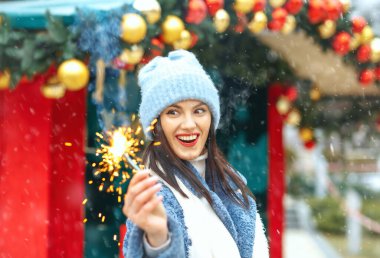 Heyecanlı genç kadın mavi ceket giyiyor. Kar yağarken Bengal ışıklarıyla tatilin tadını çıkarıyor.