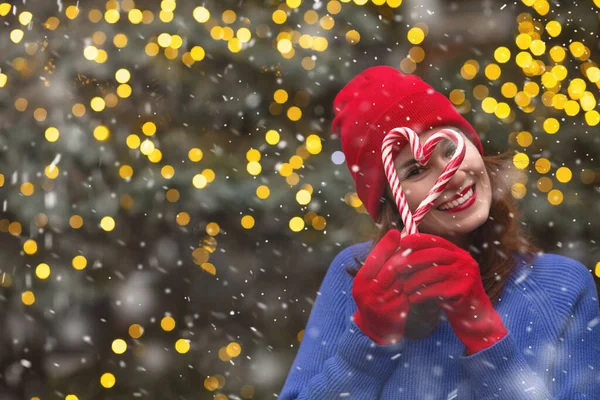 在降雪期间 一个积极的黑发女人头戴红帽子 身穿蓝色毛衣 手里拿着糖果 站在圣诞树旁 案文的篇幅 — 图库照片