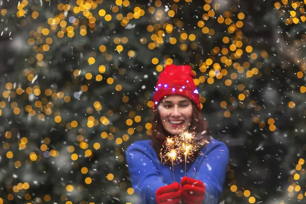 Heyecanlı esmer kadın, kar yağarken kırmızı şapka ve mavi kazak giyip Noel ağacının yanında maytaplarla eğleniyor. Metin için boşluk